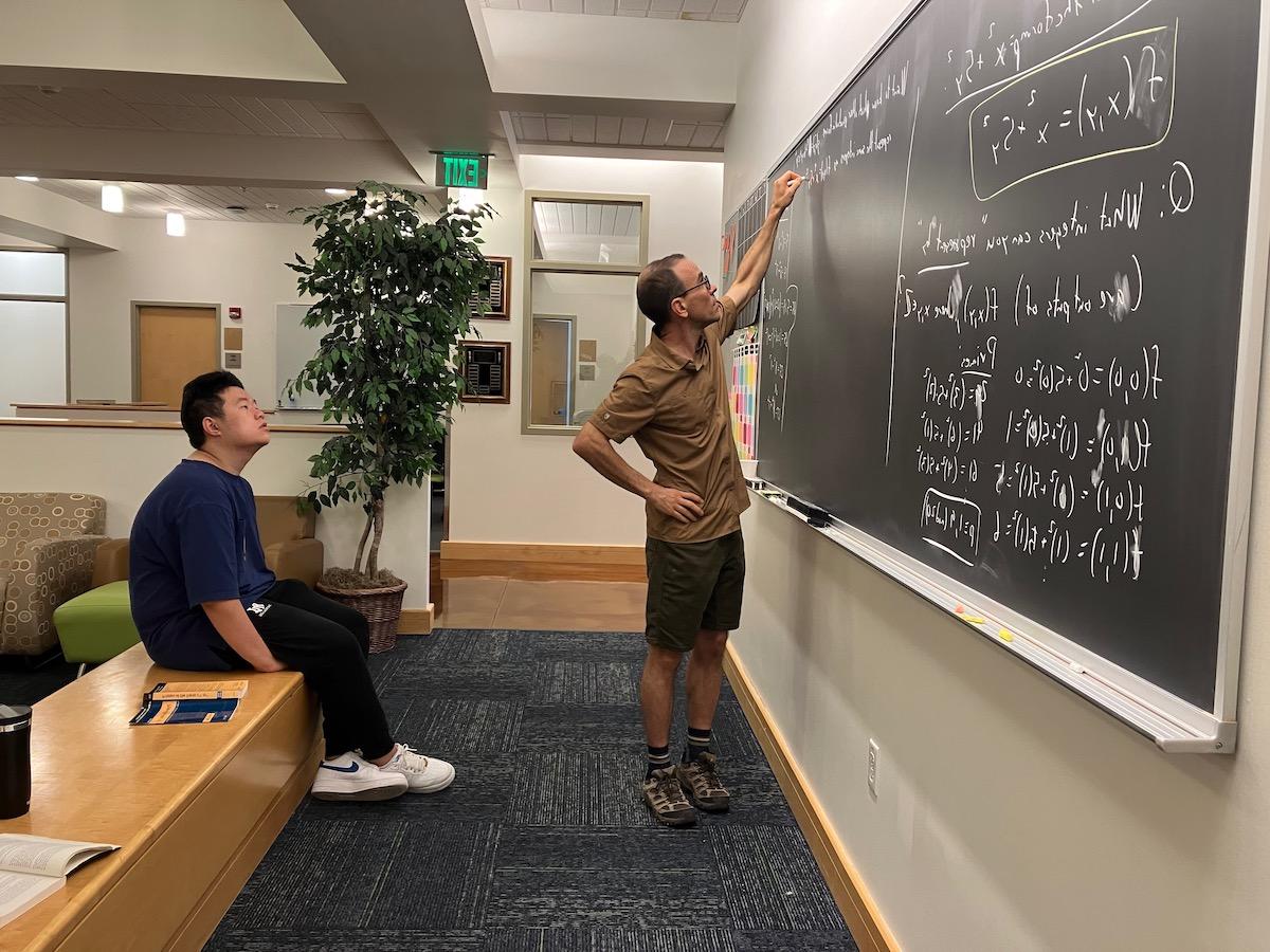 A man writing algebra functions on a whiteboard while a college student observes